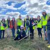 A group of students wearing white hard hats and bright safety vests stand together in a grassy field, some holding tools and clipboards, under a partly cloudy blue sky.