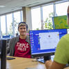 Two people sit at computer desks in the Temple Ambler library. They appear to be engaged in a conversation while working, with other library furniture visible around them.
