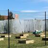 The image shows the Field Station Research Array, a large outdoor research area enclosed by a tall black mesh fence. Inside the fenced space is a grassy field with rows of evenly spaced wooden rectangular planter frames. To the right of these frames are large solar panels with plants underneath for an agrovoltaics experiment. Toward the back of the area are tall frames containing trees covered with protective netting, creating controlled research environments for a Spotted Lanternfly study.