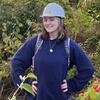 A person wearing a hard hat and backpack smiles with their hands on their hips in front of a forested background.