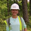 A person wearing a hard hat and safety vest smiles while standing in front of a forested background.
